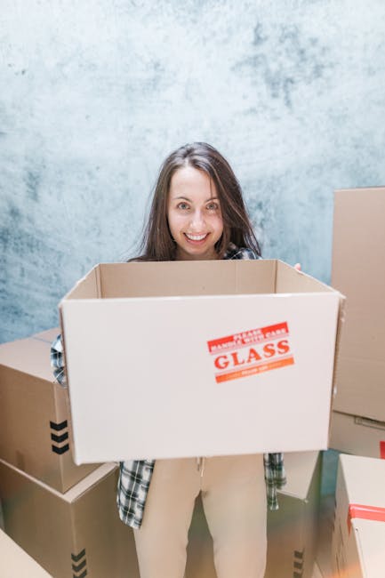Close-up of a person wearing a dark green shirt and blue pants, securing a large cardboard moving box with a white and red caution label that reads 'CAUTION THIS SIDE UP' and features an upward-pointi